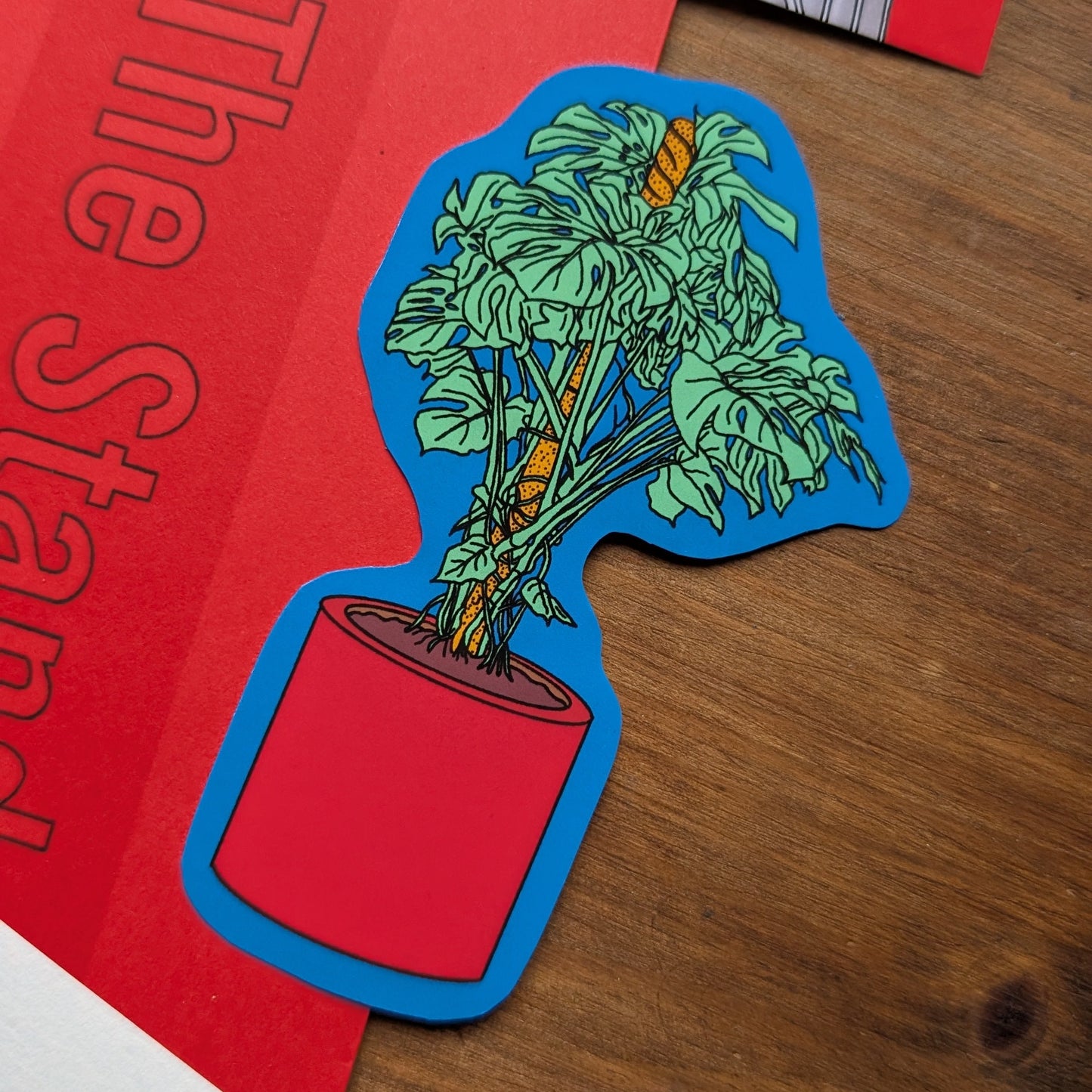 Colorful potted plant sticker on a red surface with a building in the background, formwork print club, the standard, london, monstera sticker